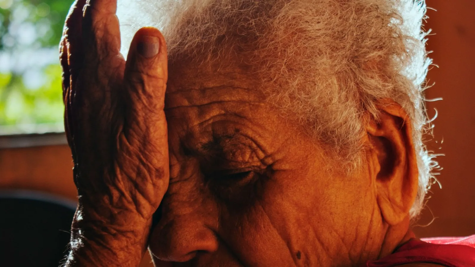 Senhora idosa sentada com a mão apoiada na testa, em ambiente doméstico com luz natural